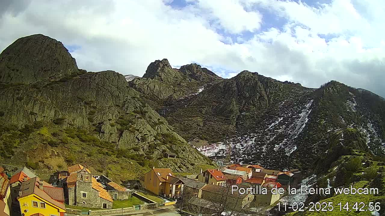 A small village nestled at the base of a rocky mountain range under a partly cloudy sky with some snow visible on higher elevations.
