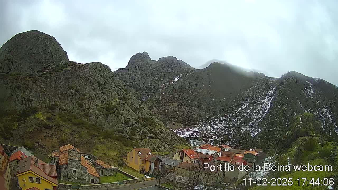 A small village nestled at the base of a large, rocky mountain range under an overcast sky with patches of snow on the higher elevations.