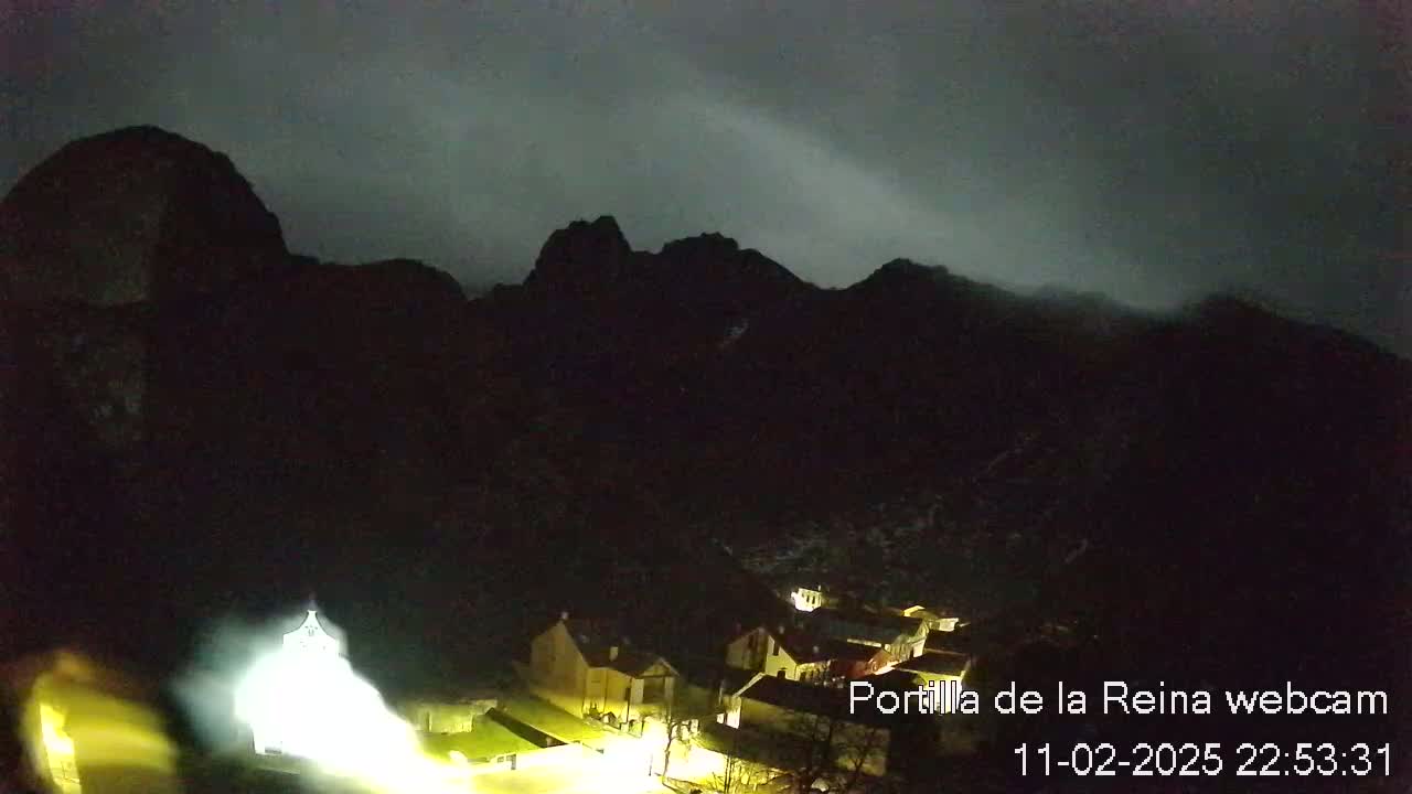 A small village nestled in a valley below dark, silhouetted mountains under a cloudy, nighttime sky.