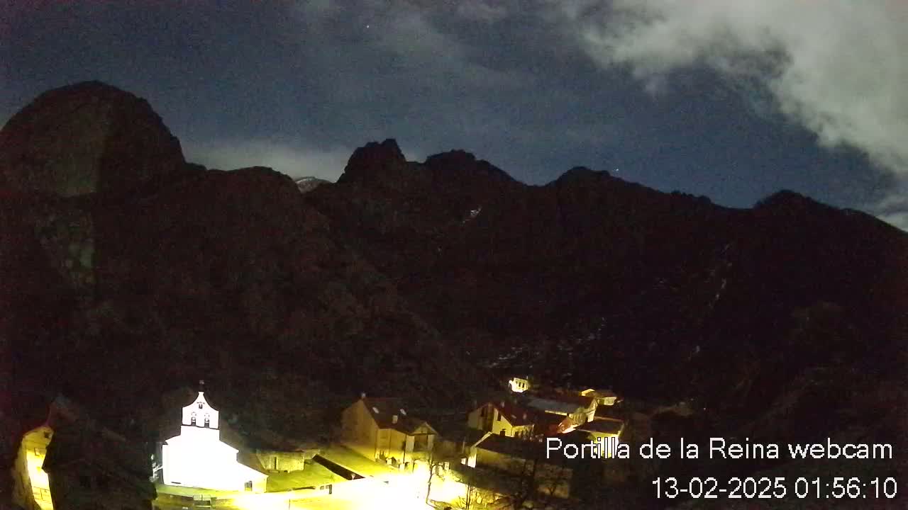 A small village nestled in a valley between dark mountains is illuminated at night under a cloudy sky.