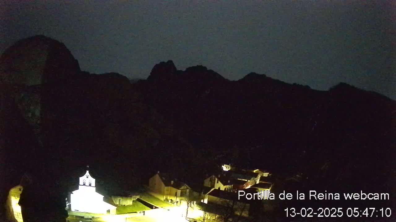 A small village illuminated at night is nestled at the base of dark mountains under a dark sky.