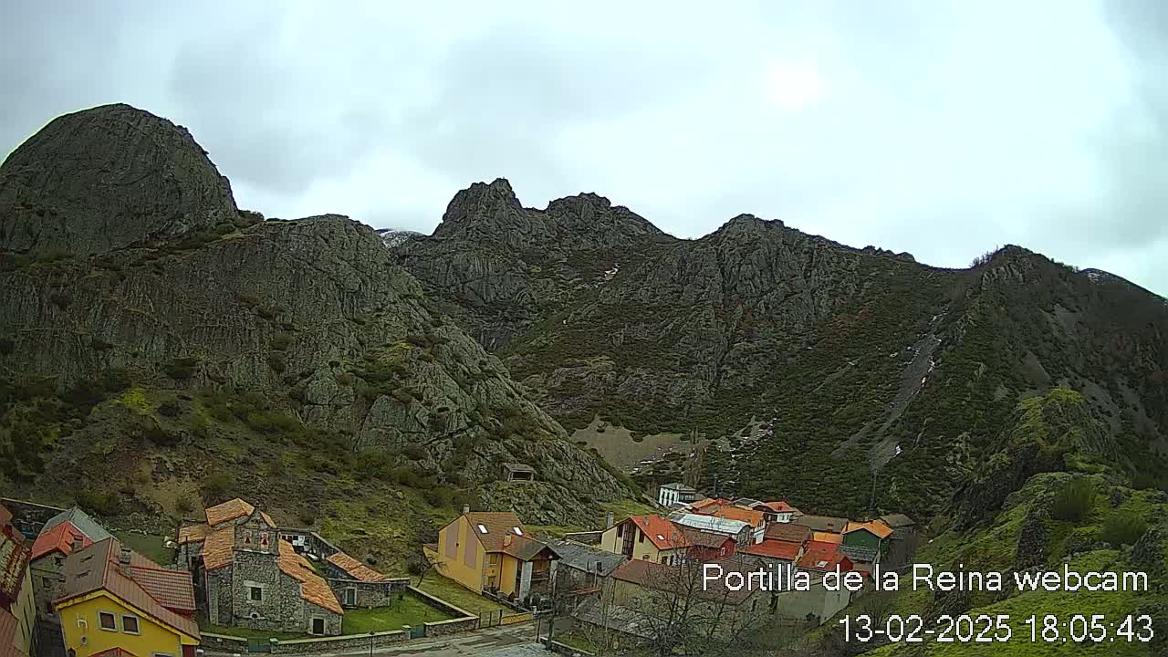 A small village nestled at the base of a large, rocky mountain range under a cloudy sky.