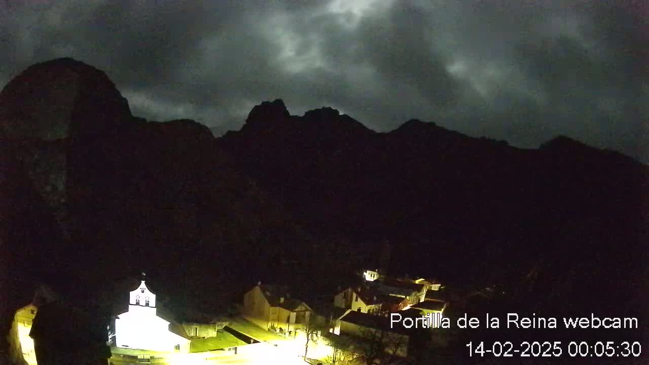 A nighttime view of a small village nestled in a valley, illuminated by artificial light, with dark, looming mountains under a cloudy sky.