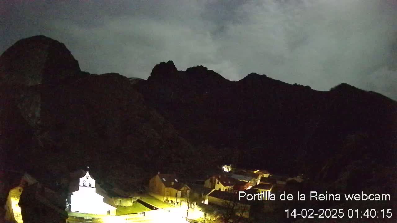 A small village nestled in a valley between dark, silhouetted mountains under a cloudy night sky.