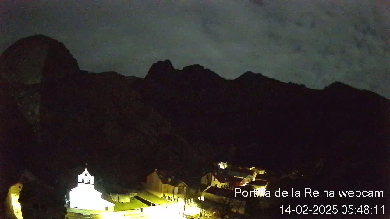 A small, illuminated village nestled at the base of dark, silhouetted mountains under a cloudy night sky.