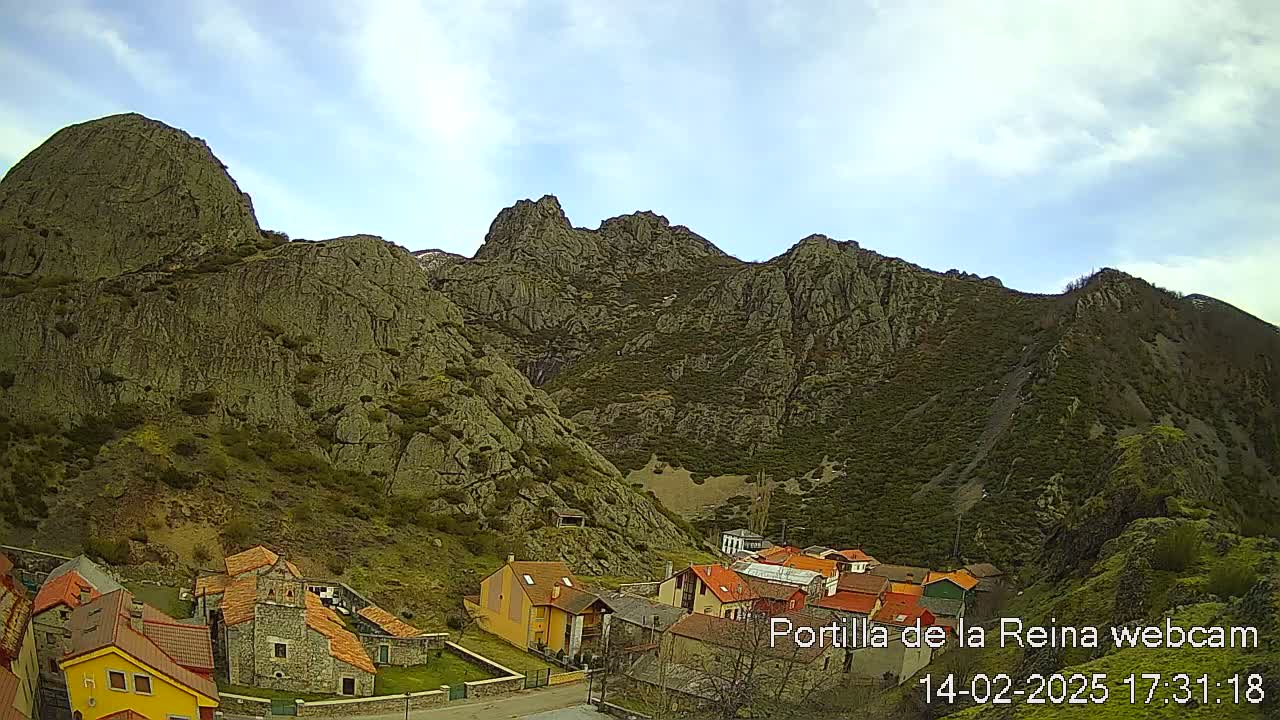 A small village with red-tiled roofs nestles at the base of a large, rocky mountain range under a partly cloudy sky.