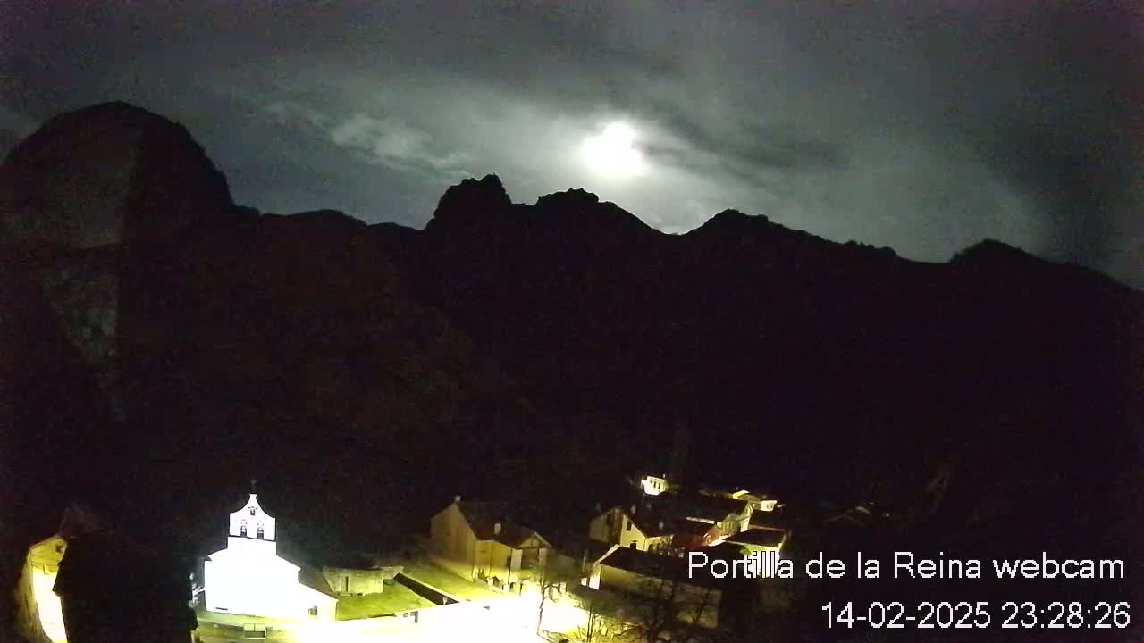 A moonlit night scene shows a small village nestled at the base of dark, mountainous terrain under a cloudy sky.