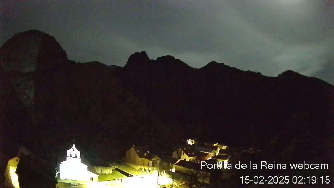 A small village nestled at the base of dark mountains is illuminated at night under a cloudy sky.