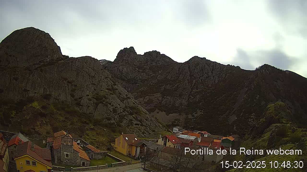 A small village nestled at the base of a large, imposing mountain range under an overcast sky.