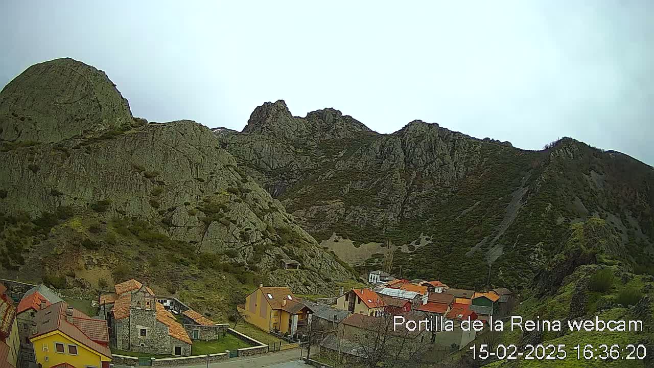 A small village nestled at the base of a large, rocky mountain range under an overcast sky.