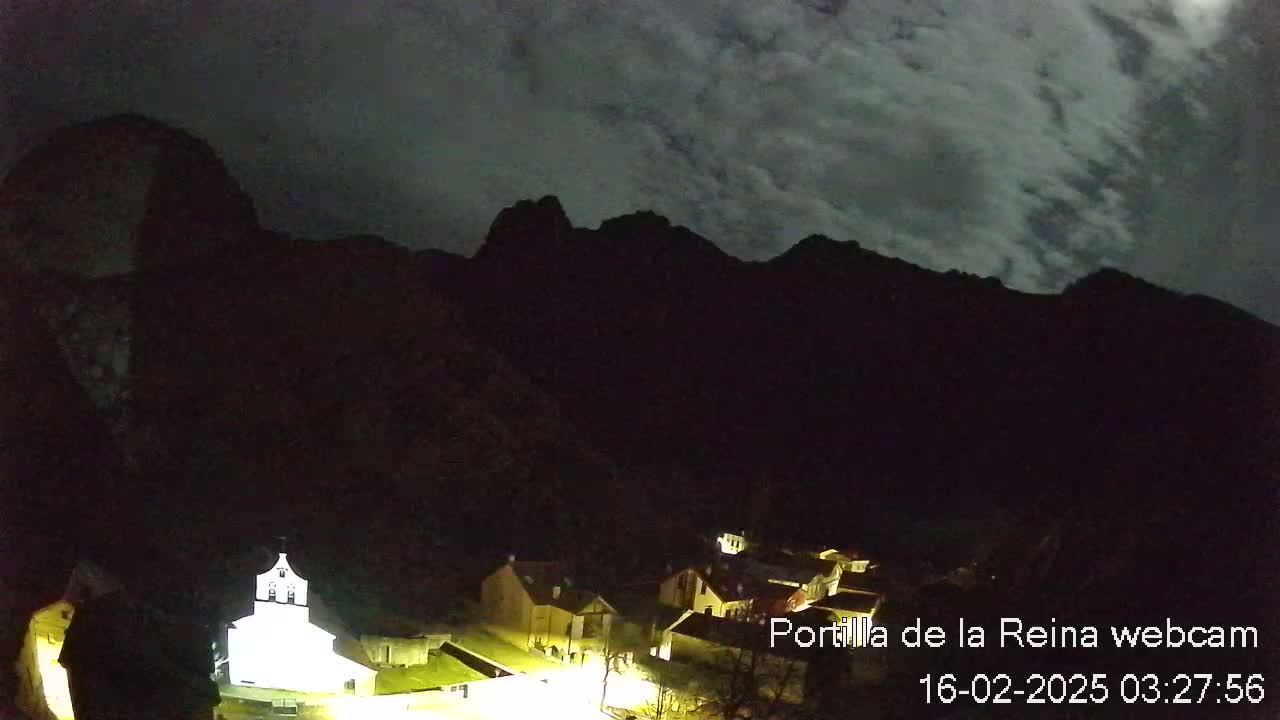 A small village nestled at the base of dark mountains under a cloudy night sky.