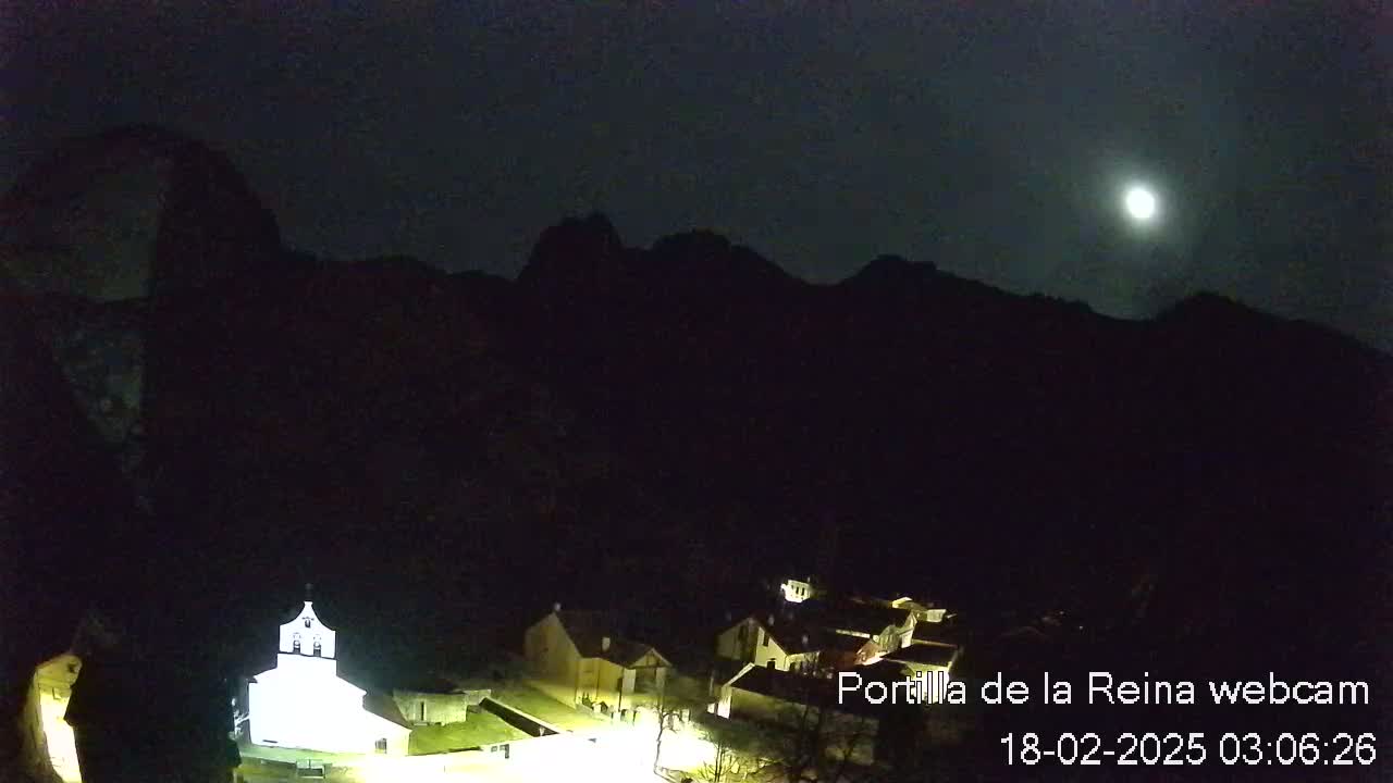 A nighttime view of a small village nestled at the base of dark mountains under a bright moon.