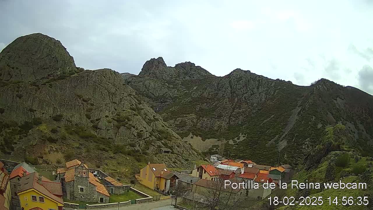 A small village nestled in a valley surrounded by rocky, green-covered mountains under a cloudy sky.