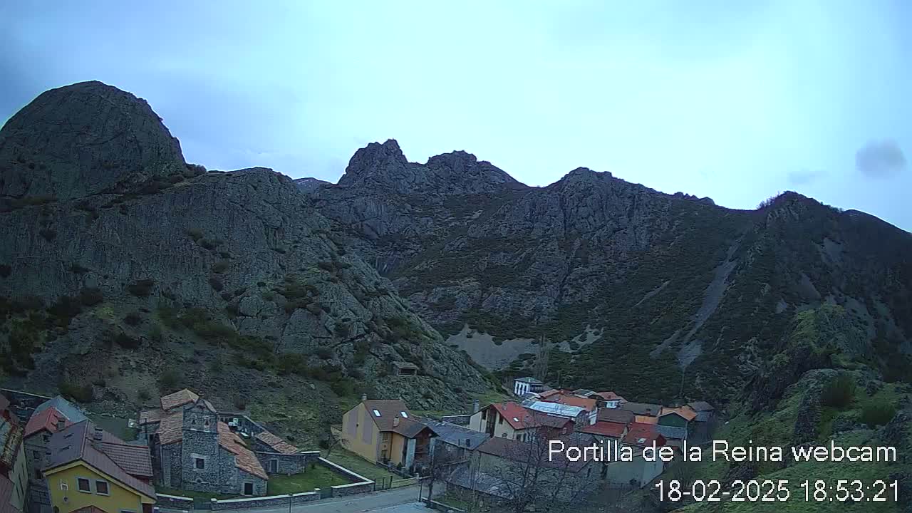 A small village nestled at the base of a large, rocky mountain range under a mostly clear, light-blue sky.