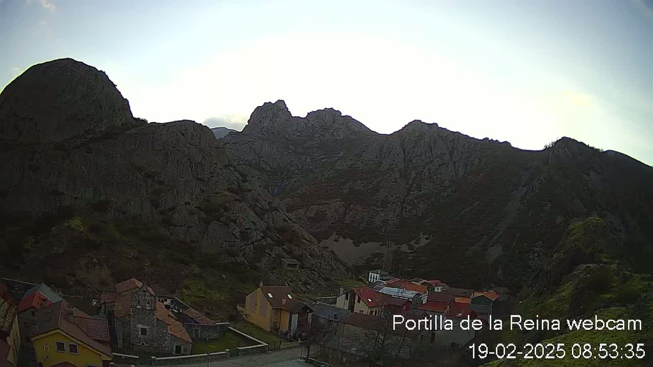A small village nestled at the base of large, dark, rocky mountains under a mostly clear sky.