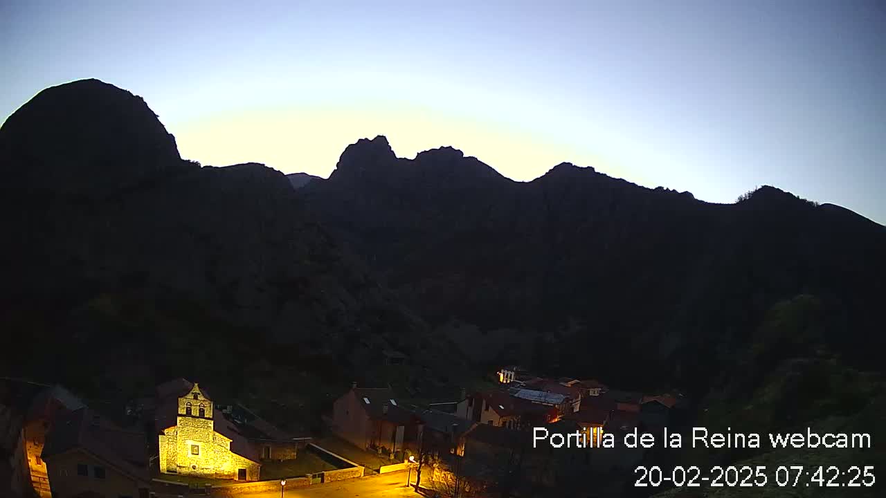 A small village nestled in a valley between dark, mountainous silhouettes is illuminated at twilight.
