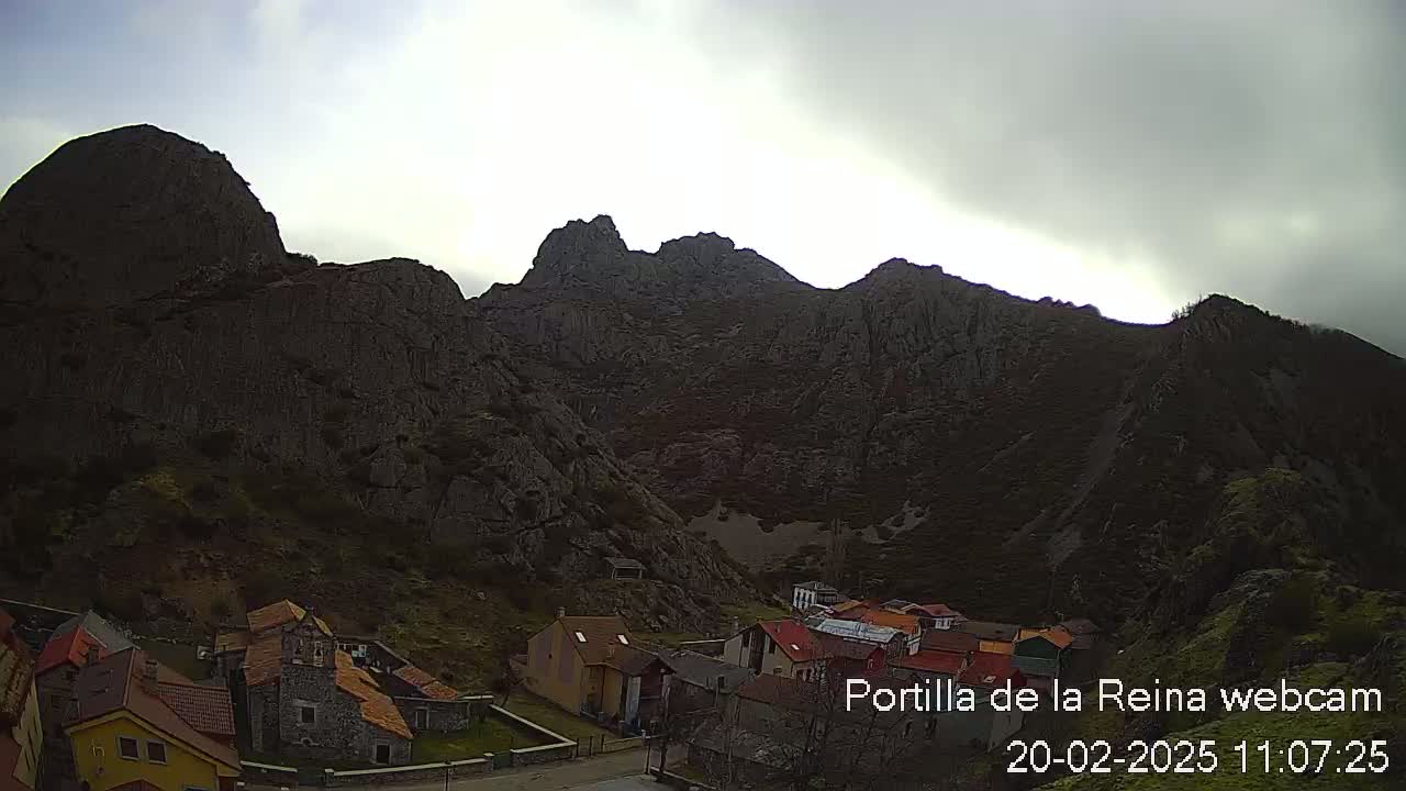A small village sits at the base of a large, dark mountain range under an overcast sky.