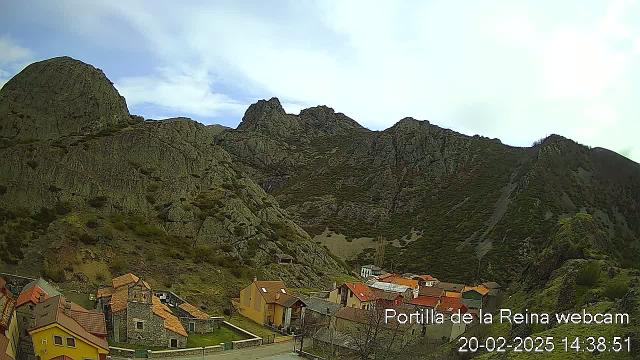 A small village nestled at the base of a large, rocky mountain range under a partly cloudy sky.
