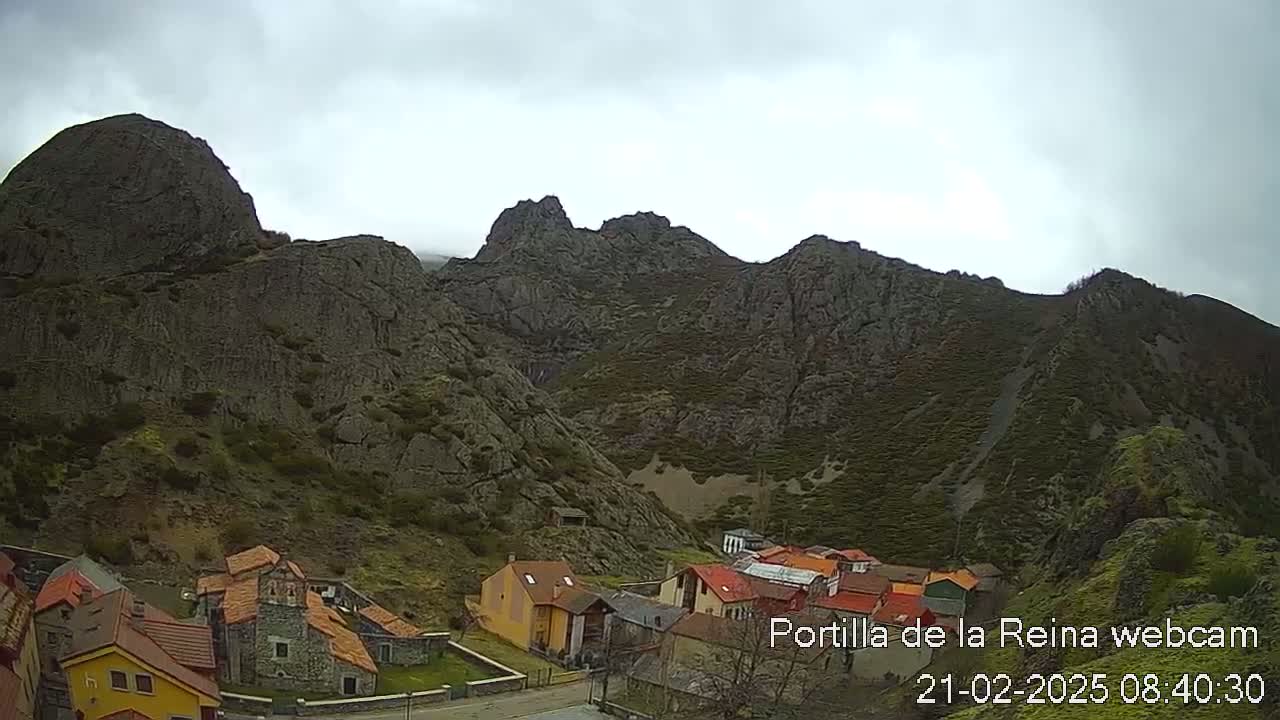 A small village nestled in a valley surrounded by imposing, dark gray rocky mountains under an overcast sky.
