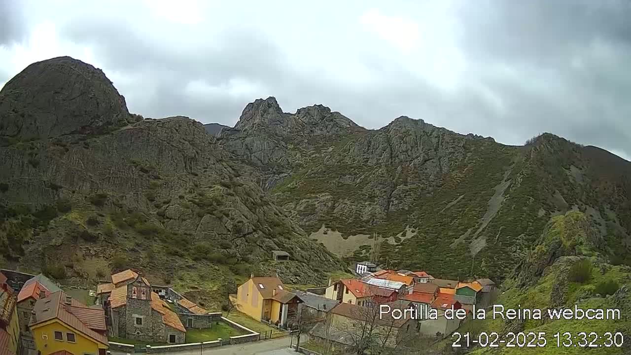 A small village of stone buildings nestles at the base of a large, rocky mountain range under an overcast sky.