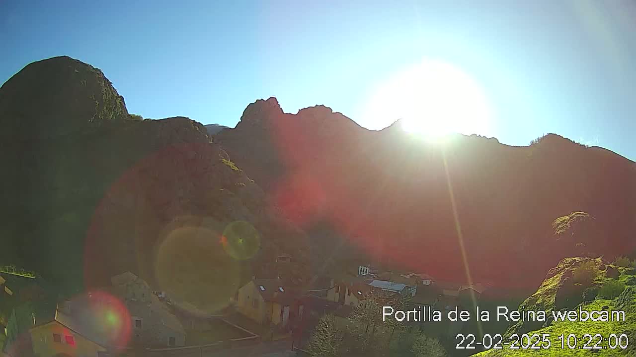 A bright sun shines over a small village nestled at the base of sunlit, rocky mountains on a clear day.