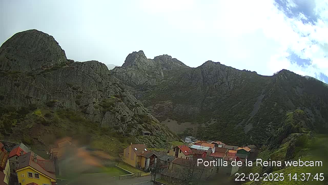 A village nestled at the base of a large, rocky mountain range under a partly cloudy sky.