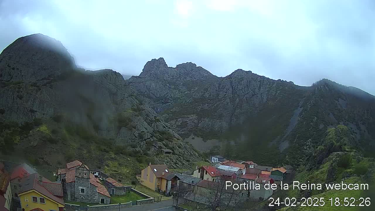 A small village nestled at the base of large, imposing, gray mountains under a cloudy sky.