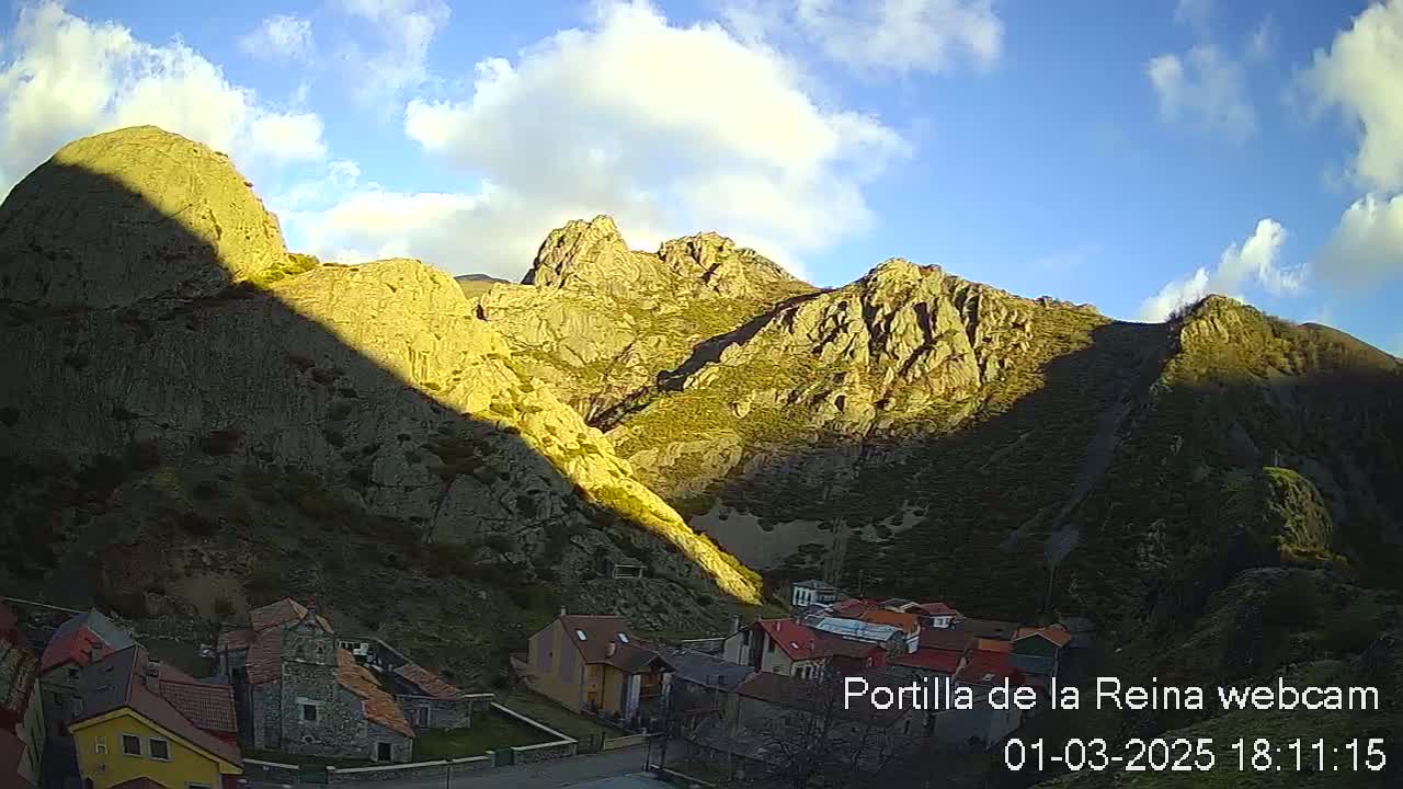 A small village nestled in a valley between sunlit, rocky mountains under a partly cloudy sky.