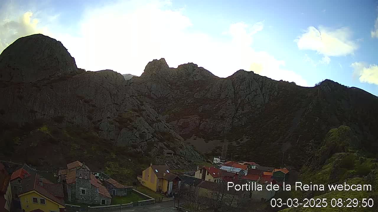 A small village nestles at the base of a large, rocky mountain range under a mostly clear, bright sky.