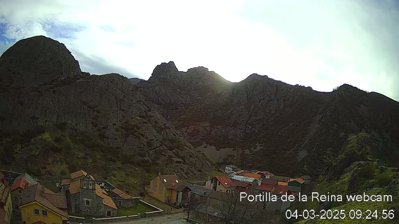 A small village nestled in a valley surrounded by large, rocky mountains under a partly cloudy sky.