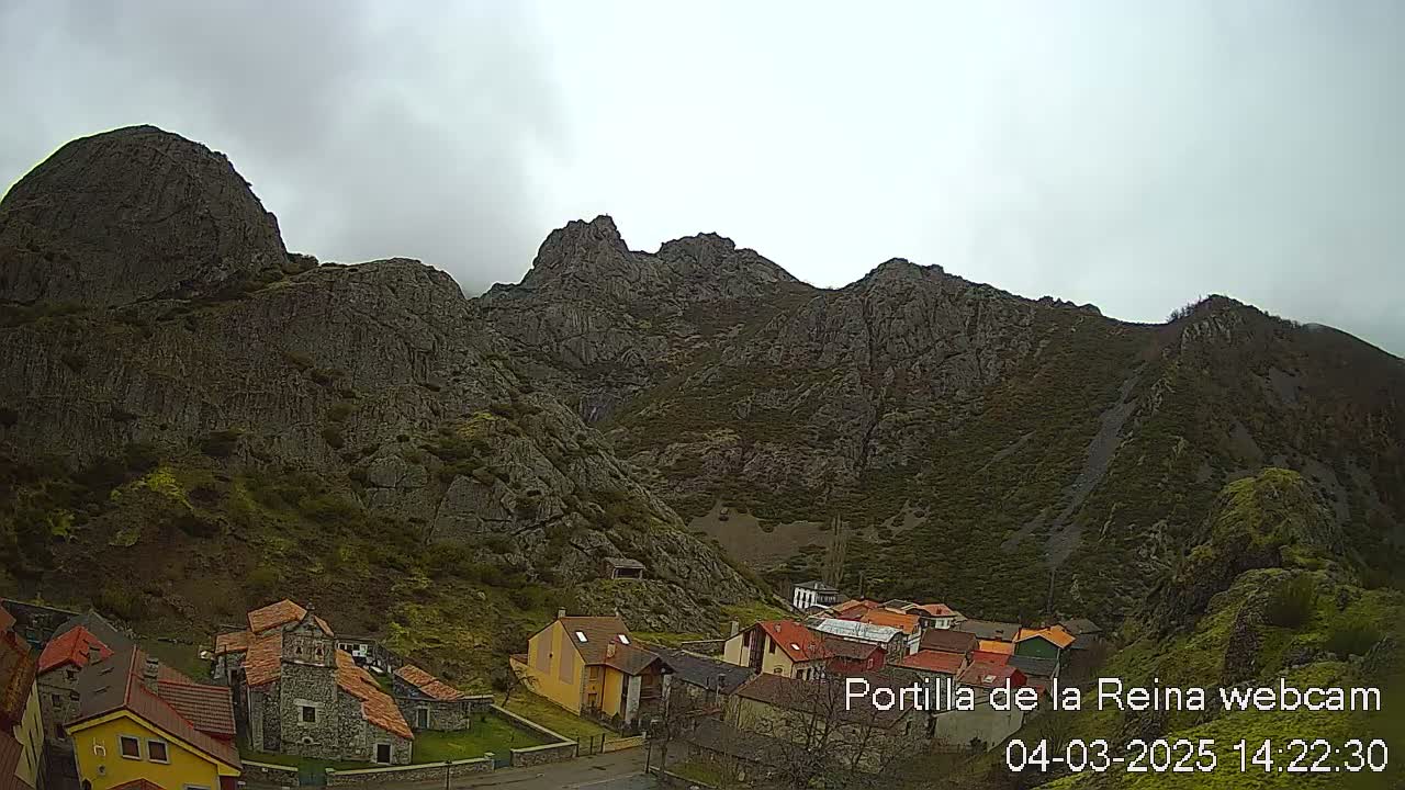 A small village nestled at the base of a large, rocky mountain range under an overcast sky.