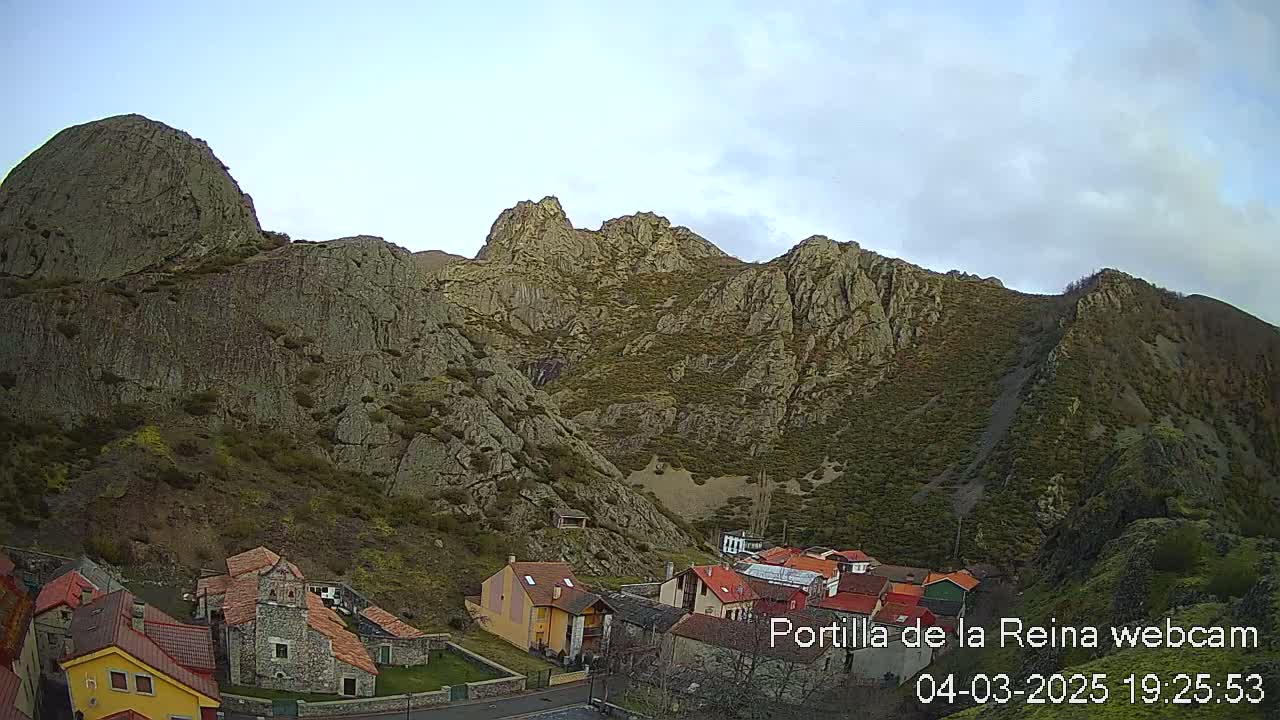 A village nestled at the base of a rocky mountain range under a partly cloudy sky.