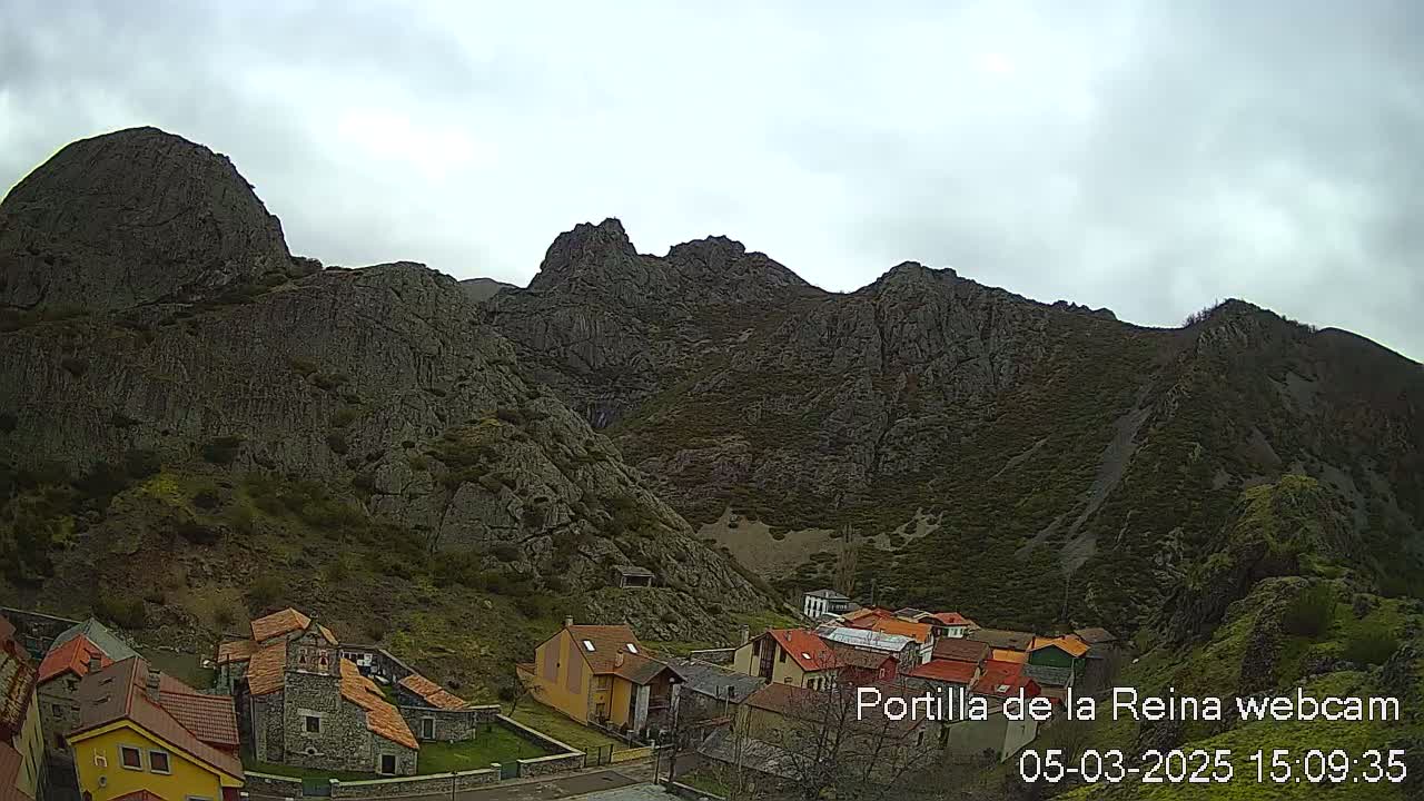 A small village nestled at the base of a large, rocky mountain range under a cloudy sky.
