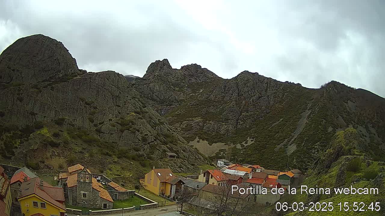 A small village nestled in a valley below a large, rocky mountain range under an overcast sky.