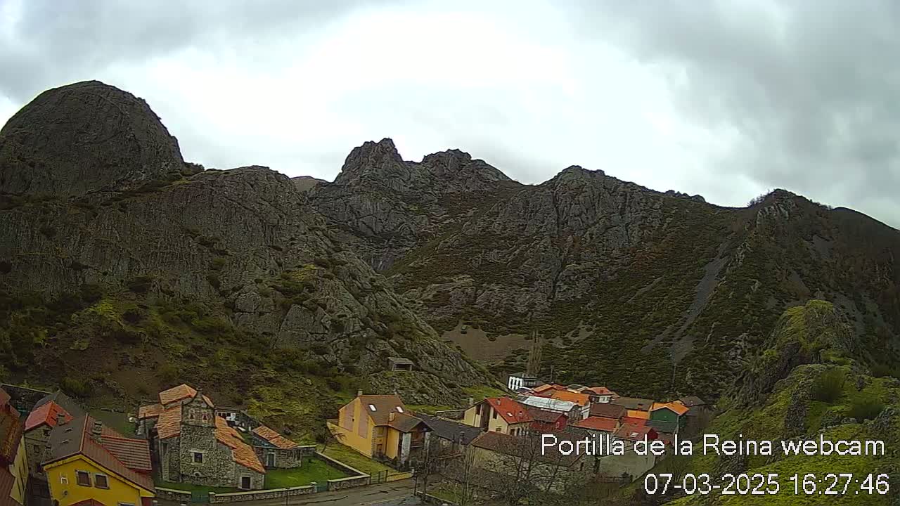 A small village nestled at the base of large, dark, rocky mountains under a cloudy sky.