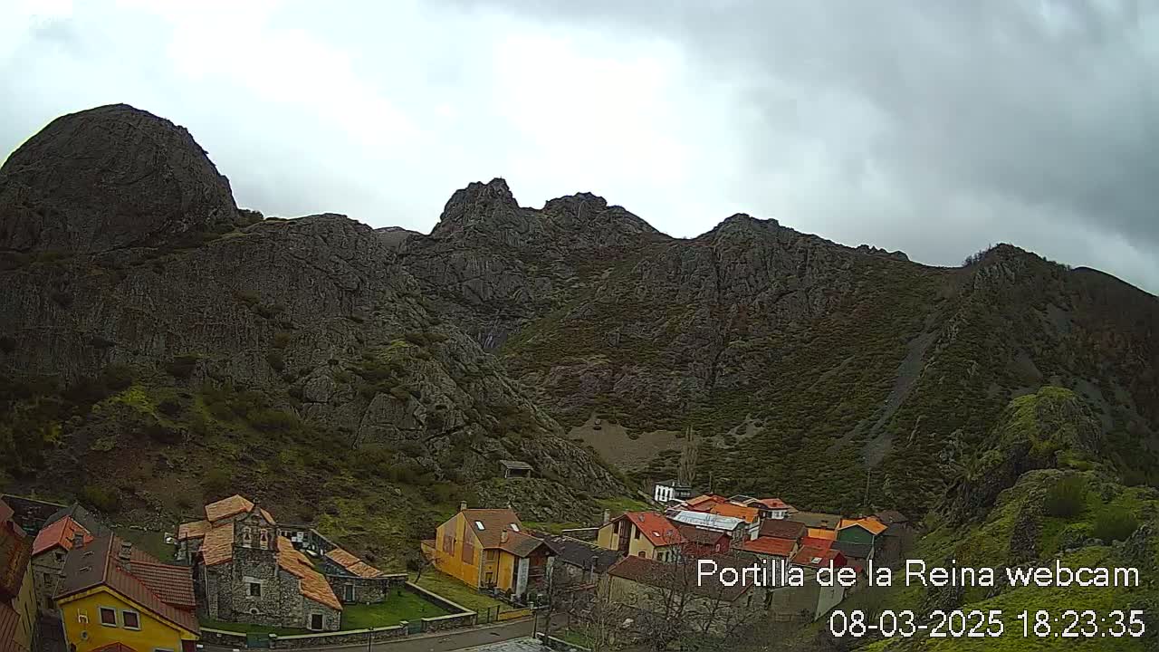 A small village sits at the base of a large, dark, rocky mountain range under an overcast sky.