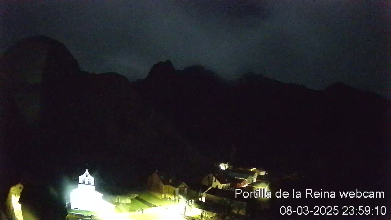 A small, illuminated village nestled at the base of dark mountains under a night sky.