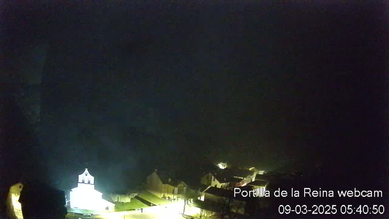 A dimly lit village is shrouded in fog at night, with a brightly lit church visible in the center.