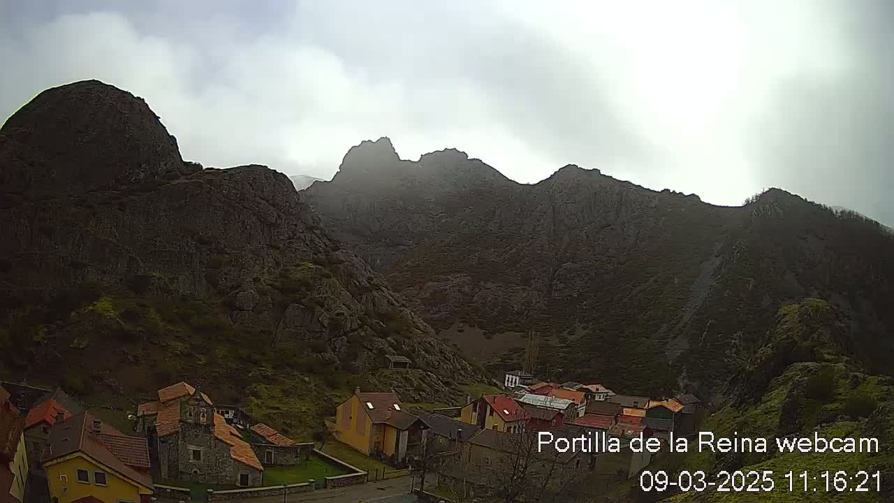 A small village nestled in a valley surrounded by imposing, mist-shrouded mountains under a cloudy sky.