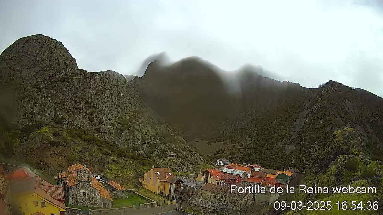 A small village nestled at the base of large, rocky mountains partially obscured by fog under an overcast sky.
