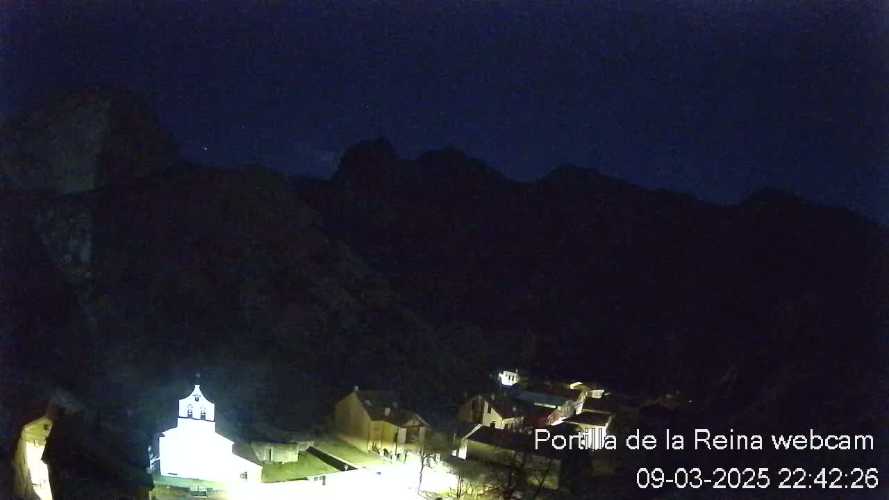 A nighttime view of a small village nestled in a dark, mountainous valley under a clear, starlit sky.