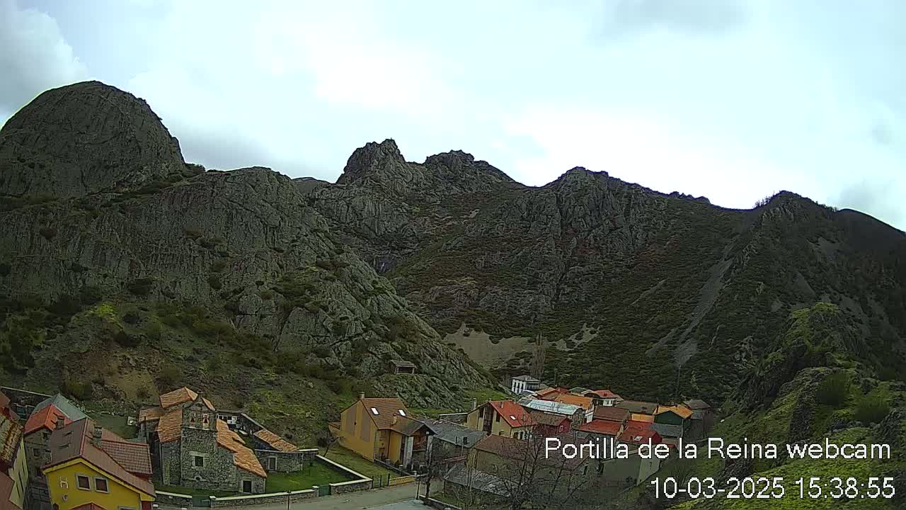 A small village nestled at the base of a large, rocky mountain range under a mostly cloudy sky.