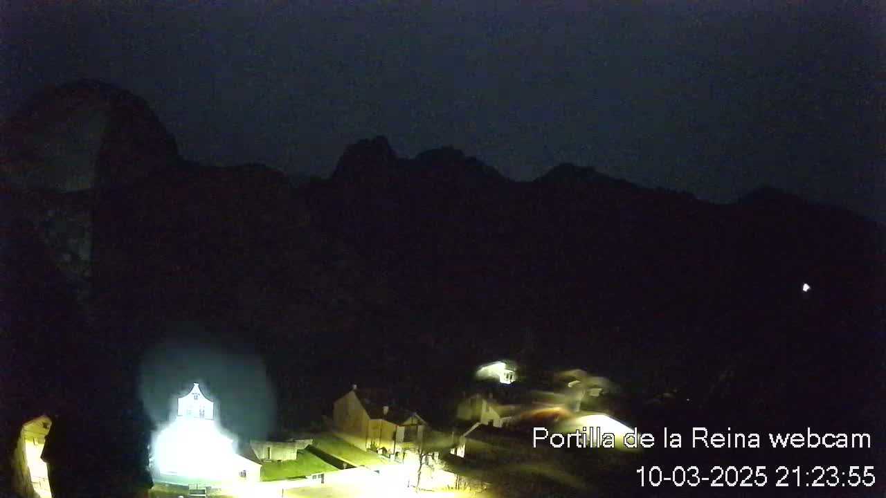 A nighttime view of a small village nestled at the base of dark mountains under a clear, dark sky.