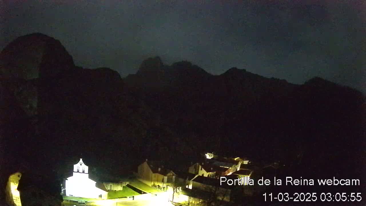 A nighttime view of a small village nestled at the base of dark mountains under a mostly clear, dark sky.