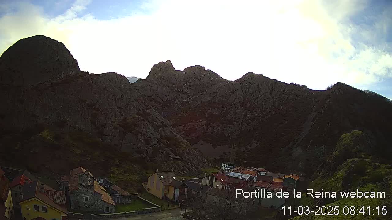 A small village nestled at the base of a large, dark mountain range under a partly cloudy sky.
