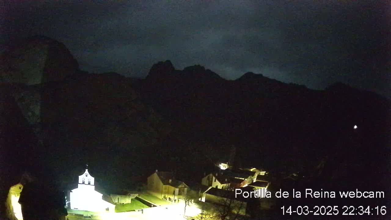 A nighttime view of a small village nestled at the base of dark, silhouetted mountains under a cloudy sky.