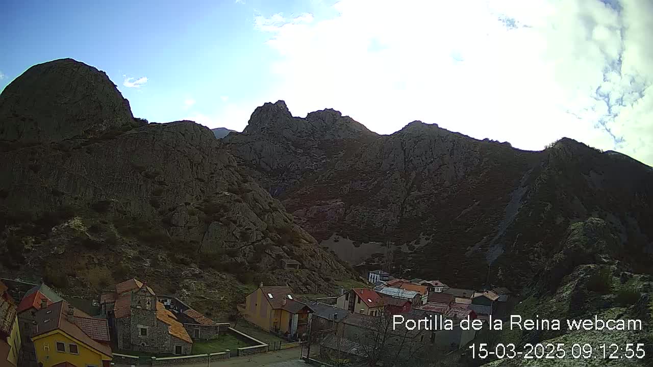 A small village nestled in a valley surrounded by rugged, dark-colored mountains under a mostly clear, bright sky.