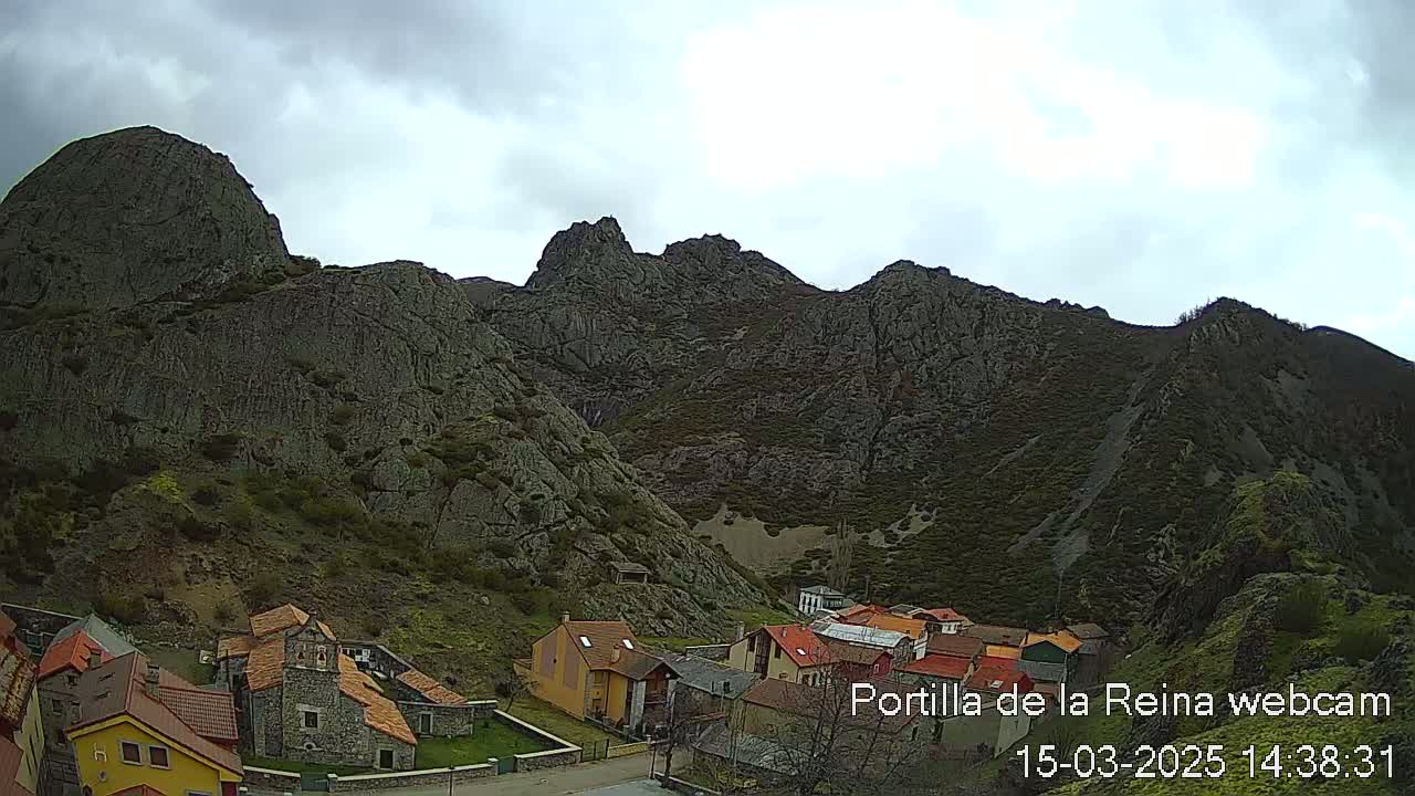 A small village nestled at the base of a large, rocky mountain range under an overcast sky.