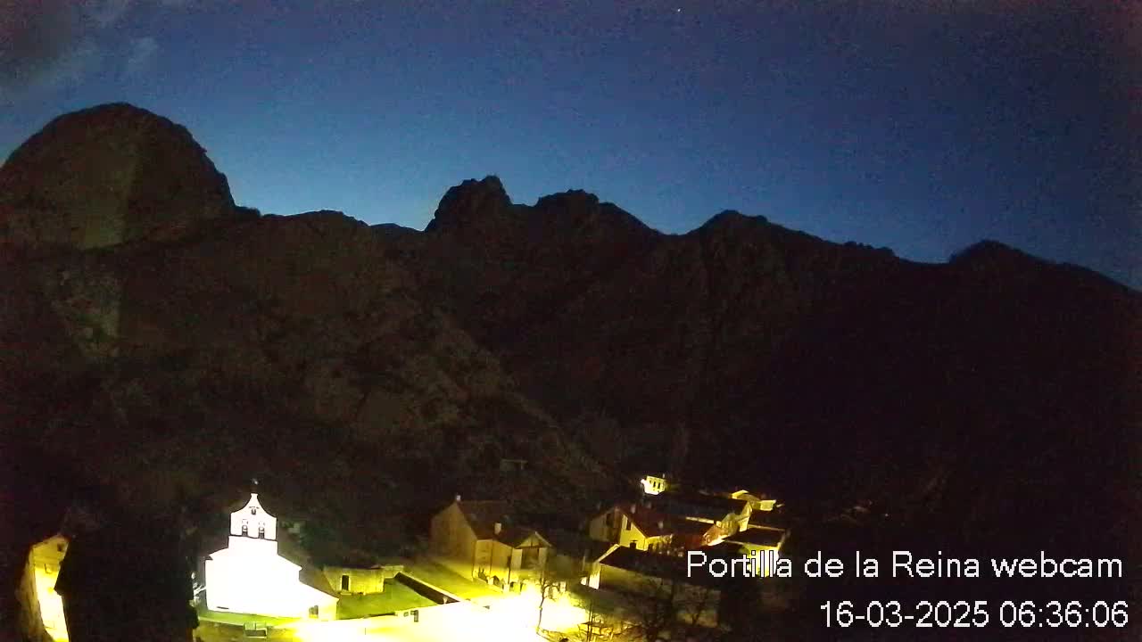 A small, illuminated village nestled at the base of dark, mountainous terrain under a twilight sky.