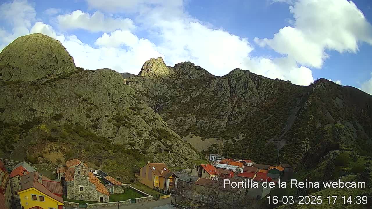 A small village nestled in a valley surrounded by large, rocky mountains under a partly cloudy sky.